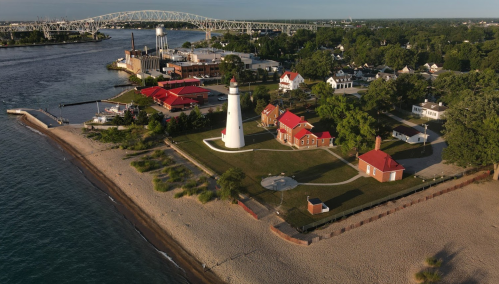 Aerial view of a lighthouse and red-roofed buildings by a sandy beach, with a bridge and river in the background.