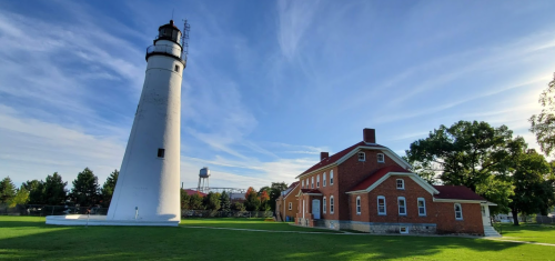 A tall white lighthouse beside a red-brick building, set against a clear blue sky and green grass.
