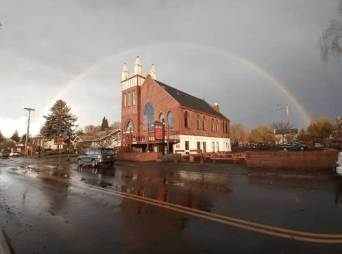 A brick building with a rainbow arching over it, reflecting on a wet street under a cloudy sky.