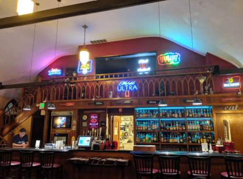 A cozy bar interior with neon signs, a wooden counter, and shelves filled with various bottles.
