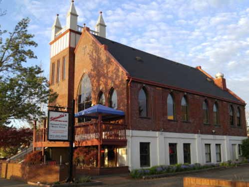 A brick building with Gothic-style windows and two spires, featuring a sign for a restaurant and outdoor seating.