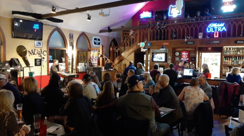 A lively bar scene with a performer on stage, patrons seated at tables, and colorful neon lights illuminating the space.