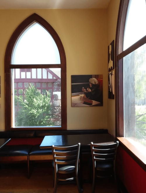 Cozy café corner with arched windows, a table, and artwork on the wall, overlooking greenery outside.