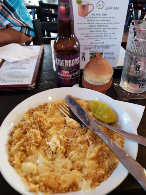 A plate of mac and cheese topped with breadcrumbs, a pickle, a roll, and a bottle of cider on a restaurant table.