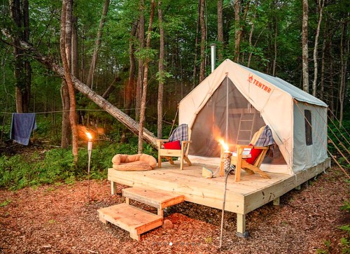 A cozy tent on a wooden platform in a forest, with chairs, a fire pit, and torches, surrounded by trees.