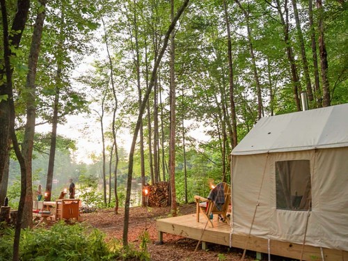 A cozy tent in a wooded area, with a wooden deck and chairs, surrounded by trees and a serene atmosphere.
