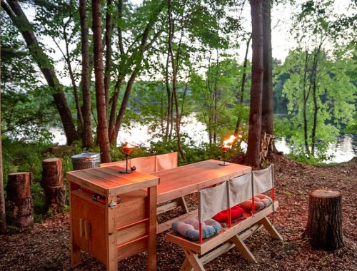 A wooden picnic table with colorful cushions, surrounded by trees near a calm lake at sunset.