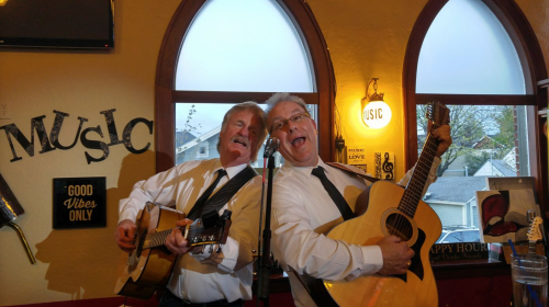 Two musicians in black ties play guitars and sing joyfully in a cozy venue with a "Music" sign on the wall.