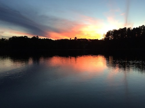 A serene sunset over a calm lake, with vibrant colors reflecting on the water and silhouettes of trees in the background.