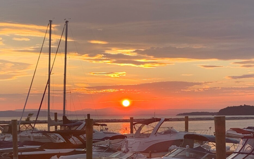 Sunset over a marina with sailboats and motorboats silhouetted against a vibrant orange and pink sky.