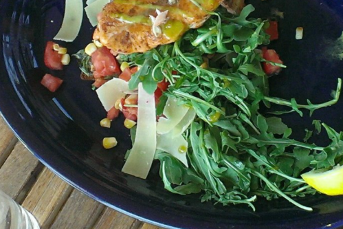 A plate of salad with arugula, tomatoes, corn, shaved cheese, and grilled salmon, garnished with lemon.