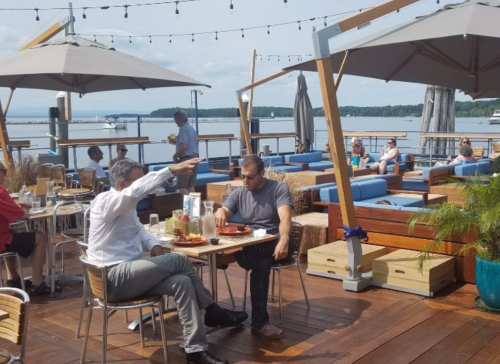 Two men dining on a waterfront patio, enjoying food and drinks, with other patrons and a scenic view in the background.