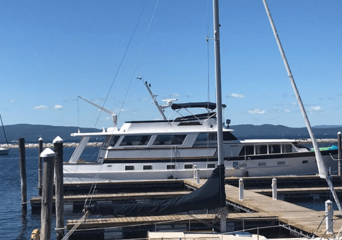 A large yacht docked at a marina on a sunny day, with blue skies and distant mountains in the background.