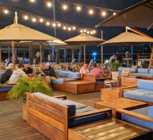 A lively outdoor seating area at night, featuring wooden furniture, umbrellas, and string lights, with people dining and socializing.