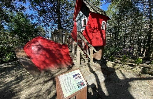 A whimsical red house on a wooden platform surrounded by trees, with an informational sign in the foreground.