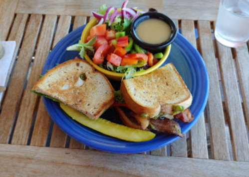 A plate with a sandwich, a side salad in a bowl, and a pickle on a wooden table.