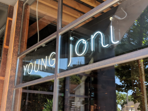 Neon sign reading "YOUNG joni" in a restaurant window with wooden framing and greenery outside.