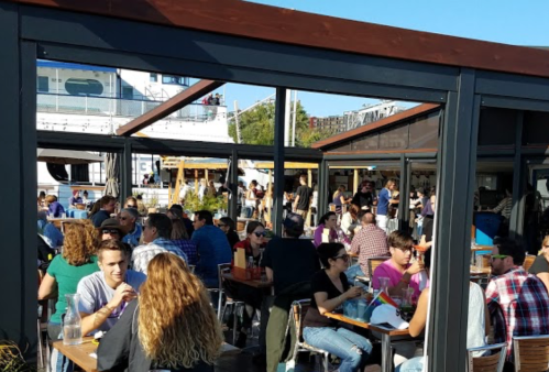A bustling outdoor dining area filled with people enjoying food and drinks under a clear blue sky.