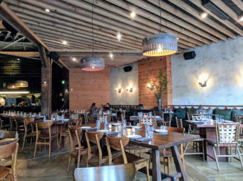 A spacious restaurant interior with wooden beams, tables set for dining, and modern light fixtures.