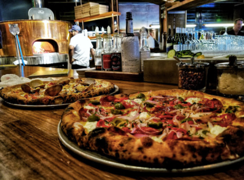 Two pizzas on a wooden table, with a brick oven in the background and drinks lined up behind them.
