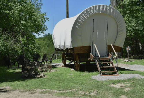 A covered wagon on a grassy area, with wooden steps and chairs nearby, surrounded by trees and a clear blue sky.