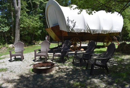 A covered wagon sits near a fire pit, surrounded by wooden chairs in a wooded area.