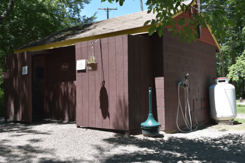 A small brown shed with a red roof, surrounded by trees and gravel, featuring a propane tank and water hose.