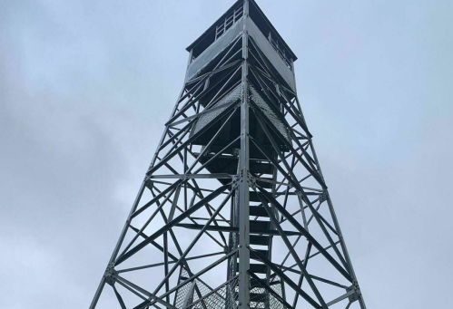 A tall metal observation tower against a cloudy sky, featuring a staircase and a small cabin at the top.