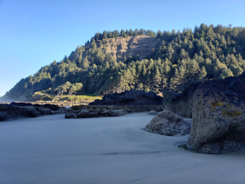 A serene beach scene with rocky formations, sandy shore, and a forested hillside under a clear blue sky.