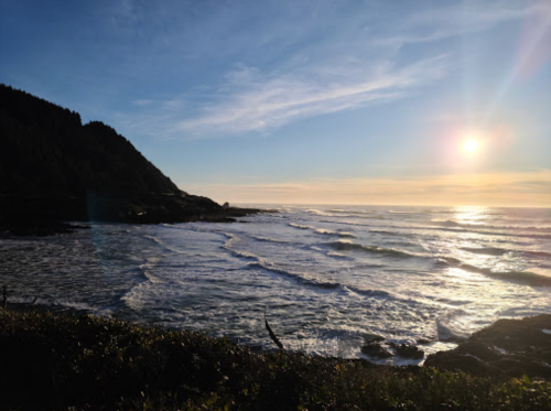 A serene coastal scene at sunset, with gentle waves and a sun setting behind a distant hill.
