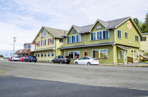 A green two-story building with large windows, parked cars, and a street view under a clear sky.