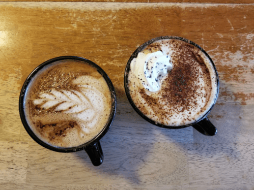 Two coffee cups on a wooden table: one with latte art and the other topped with whipped cream and cocoa powder.