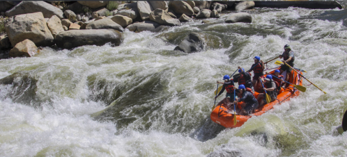 A group of people in an orange raft navigating through turbulent whitewater rapids surrounded by rocks.