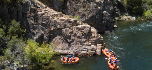 A group of people in orange rafts navigating a river near rocky cliffs and lush greenery.