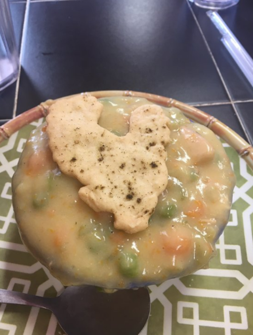 A bowl of creamy vegetable soup topped with a chicken-shaped cracker, resting on a decorative placemat.
