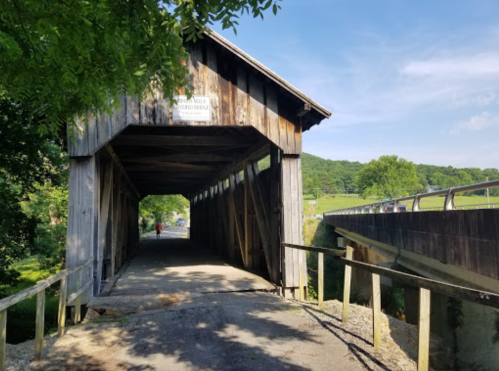 A wooden covered bridge with a gravel path, surrounded by greenery and a modern bridge in the background.