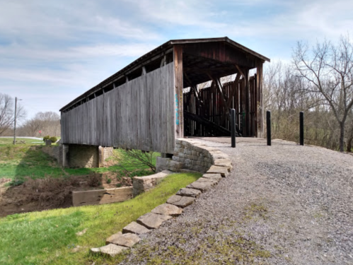 A wooden covered bridge spans a small creek, surrounded by grassy areas and trees under a cloudy sky.
