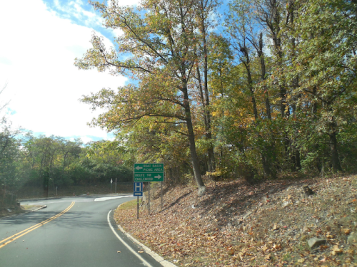 A winding road with a green directional sign for "Exit Barn" and "Bullet Two," surrounded by autumn trees.