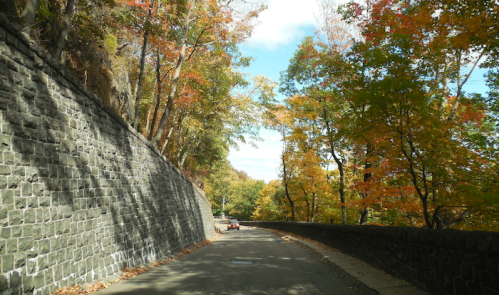 A scenic road lined with colorful autumn trees and a stone wall on one side, under a partly cloudy sky.