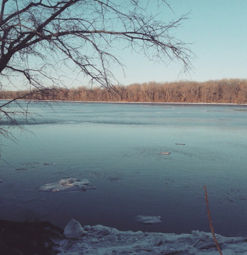 A serene winter scene of a river with ice patches, framed by bare trees against a clear blue sky.