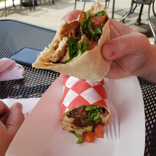 A hand holding a half-eaten wrap filled with chicken, lettuce, and tomato, next to a whole wrap in a red-checkered paper.