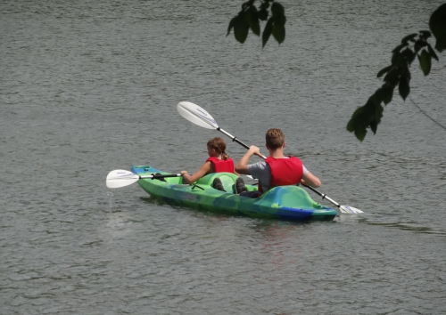 Two people in a kayak on a calm lake, wearing red life jackets and paddling together.