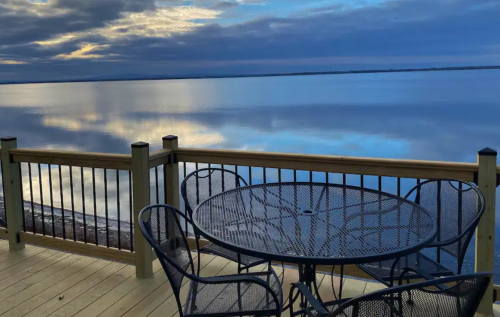 A serene lakeside view at dusk, featuring a table and chairs on a wooden deck overlooking calm waters.