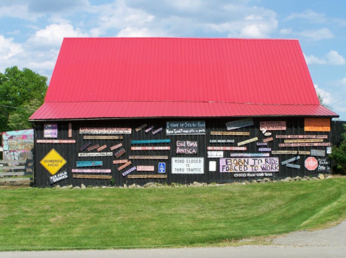 A black barn with a red roof covered in various colorful signs and messages, set against a blue sky.