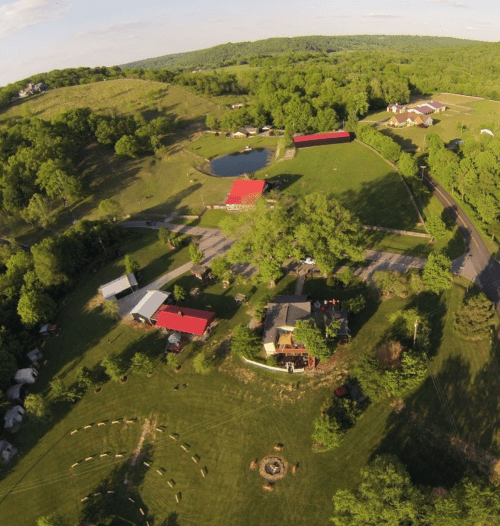 Aerial view of a green landscape with buildings, a pond, and a circular fire pit surrounded by trees.
