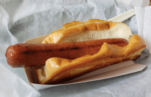 A hot dog in a bun, served in a paper tray, resting on a light-colored surface.