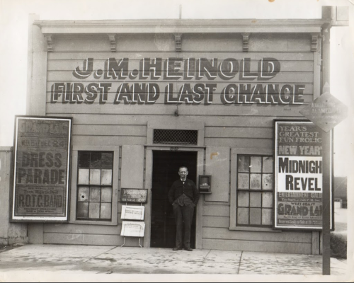 A historic building with signs reading "J.M. Heinold First and Last Chance," featuring a man standing at the entrance.