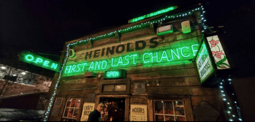A rustic bar with neon signs reading "Heinold's First and Last Chance" and "Open," adorned with festive lights at night.