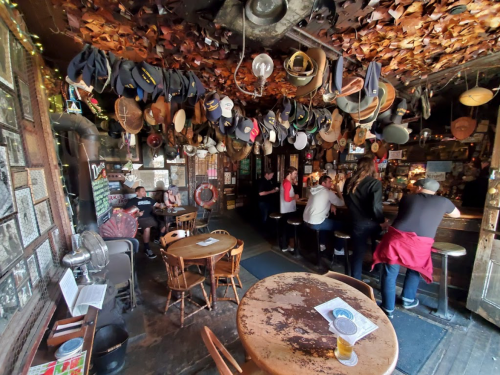 A cozy bar with a ceiling covered in hats, wooden tables, and patrons enjoying drinks and conversation.