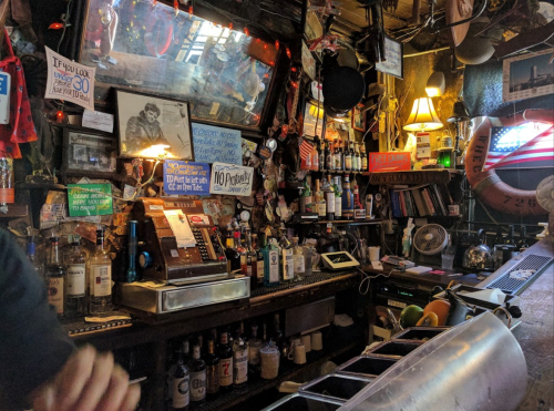 A cluttered bar with various bottles, signs, and decorations, featuring a cash register and a fan in a cozy atmosphere.